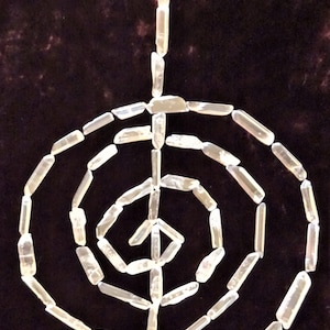 May include: A white crystal quartz labyrinth, also known as a crystal grid, is arranged in a spiral pattern on a dark brown velvet background. The crystal points are all facing the same direction, creating a sense of movement and energy.