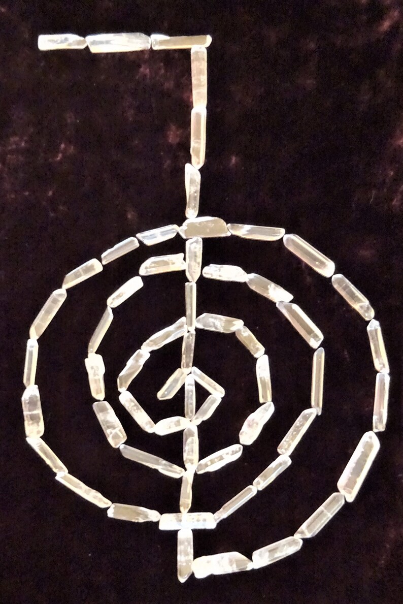 May include: A white crystal quartz labyrinth, also known as a crystal grid, is arranged in a spiral pattern on a dark brown velvet background. The crystal points are all facing the same direction, creating a sense of movement and energy.