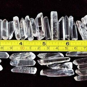 May include: A collection of approximately 40 clear quartz crystal points, ranging in size from about 1 to 2 inches long. The crystals are arranged in two rows on a dark red surface, with a yellow measuring tape placed between the rows. The tape measures up to 10 feet.