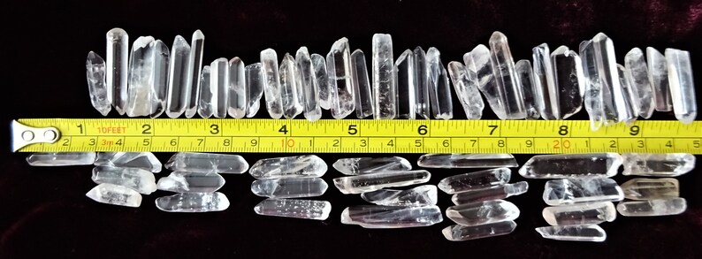 May include: A collection of approximately 40 clear quartz crystal points, ranging in size from about 1 to 2 inches long. The crystals are arranged in two rows on a dark red surface, with a yellow measuring tape placed between the rows. The tape measures up to 10 feet.