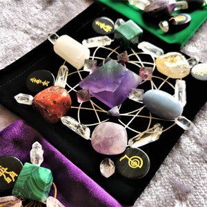 May include: A black velvet pouch with a grid pattern and various crystals and stones arranged in a circle. The crystals include amethyst, clear quartz, red jasper, and green aventurine. The pouch also has black circular stones with gold symbols.