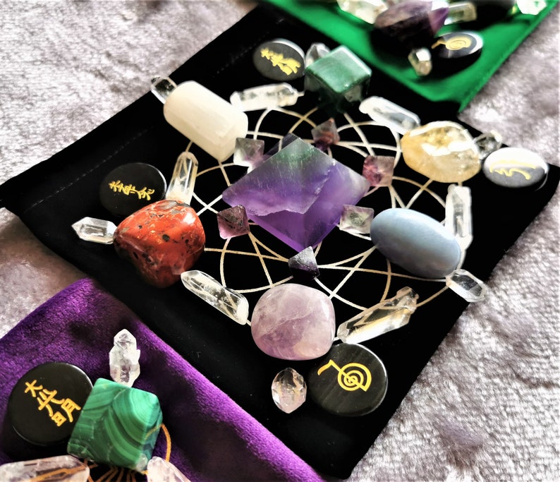May include: A black velvet pouch with a grid pattern and various crystals and stones arranged in a circle. The crystals include amethyst, clear quartz, red jasper, and green aventurine. The pouch also has black circular stones with gold symbols.