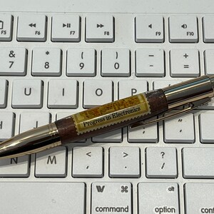 May include: A gold-toned pen with a wooden barrel and a clip, resting on a white keyboard. The pen features the text "Progress in Electronics" on a rectangular label. The pen's design includes a gold tip and accents.