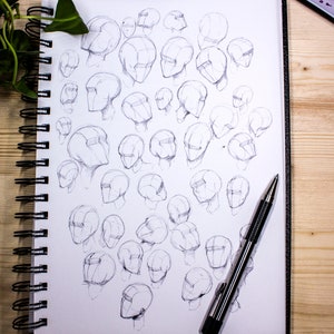 May include: A sketchbook filled with pencil drawings of human heads in various angles and positions. A silver pen is lying on top of the sketchbook.