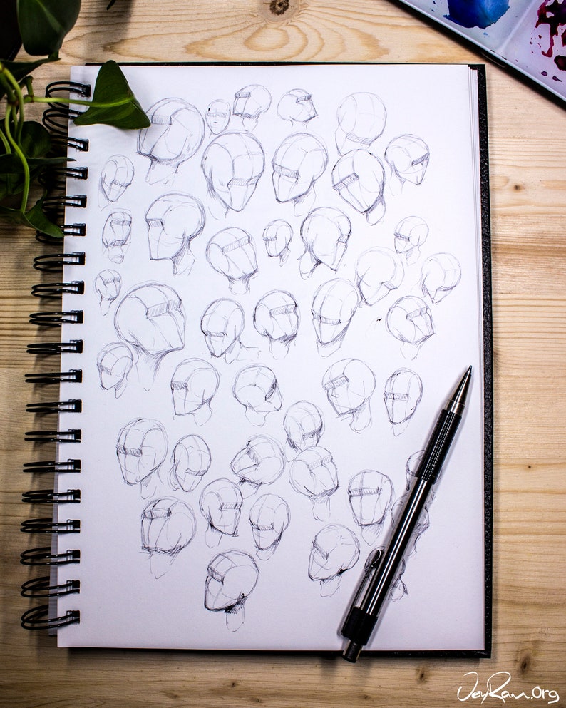 May include: A sketchbook filled with pencil drawings of human heads in various angles and positions. A silver pen is lying on top of the sketchbook.
