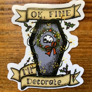 May include: A white sticker with a black and gray coffin decorated with a red bow and a wreath. The coffin is surrounded by green vines and holly. The sticker has two banners with the text "OK, FINE I'll Decorate."