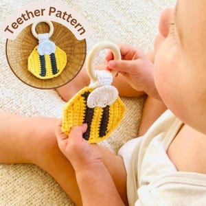 May include: A handmade crochet baby teether in the shape of a bee. The teether features a yellow body with black stripes and white wings. The text "Teether Pattern" is in the upper left corner. The teether is held by a baby.