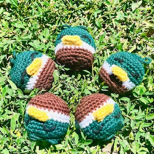 May include: Five handmade crochet ducklings in shades of green, brown, white, and yellow, arranged on a bed of green grass. Each duckling features a detailed design, with a yellow beak and a white stripe. The ducklings are approximately 2 inches in diameter.