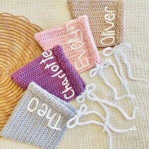 May include: Five crocheted pouches in various colors: tan, pink, purple, and gray. Each pouch has a name embroidered in white: "Oliver", "Evelyn", "Charlotte", and "Theo". White drawstrings with bows are attached to the pouches.