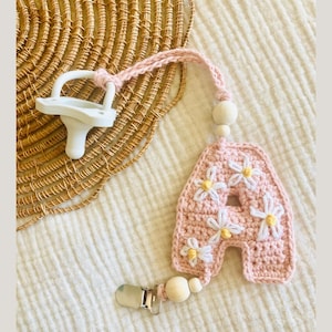 May include: A pink crocheted letter A with white daisy flowers, attached to a wooden bead and a silver clip. The letter is a baby pacifier clip.