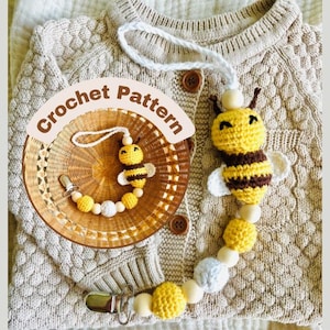 Könnte beinhalten: Ein handgefertigter Schnullerhalter mit gehäkelter Biene mit gelben, braunen und weißen Akzenten. Der Clip hat ein Bienen-Design und Holzperlen. Die Worte "Crochet Pattern" sind in einem kreisförmigen Design.
