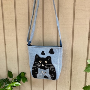 以下が含まれることがあります： 黒い猫と子猫のアップリケが付いた青いデニムのクロスボディバッグ。猫と子猫はハートを持っています。