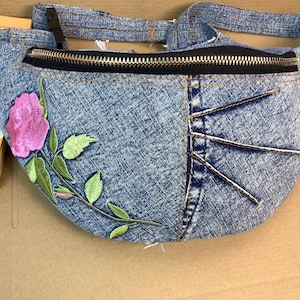 May include: A denim fanny pack with a zipper closure and a floral embroidery design. The embroidery features a pink rose and green leaves.