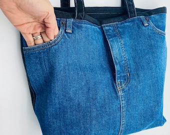 May include: A blue denim tote bag with black straps. The bag has a front pocket and a seam down the centre.