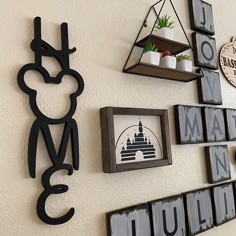 May include: Black wooden home decor sign with the word "HOME" spelled out in a stylized font. The "O" is a Mickey Mouse head. The sign is hanging on a white wall with other decor, including a framed print of the Cinderella Castle silhouette and wooden tiles with letters spelling out "JOY" and "MATTHEW".