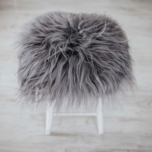 May include: A grey fluffy sheepskin rug sits on a white stool. The rug is soft and plush, and would be perfect for adding a touch of warmth and comfort to any room.