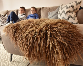Op de afbeelding: Een bruin nep bontkleed met lange, ruwe vezels. Het kleed is gedrapeerd over een lichtbruine bank met kussens.