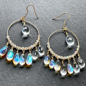 May include: A pair of gold hoop earrings with clear and iridescent teardrop-shaped crystals. The earrings have a delicate design with small clear beads and gold hooks. The crystals reflect a rainbow of colors.