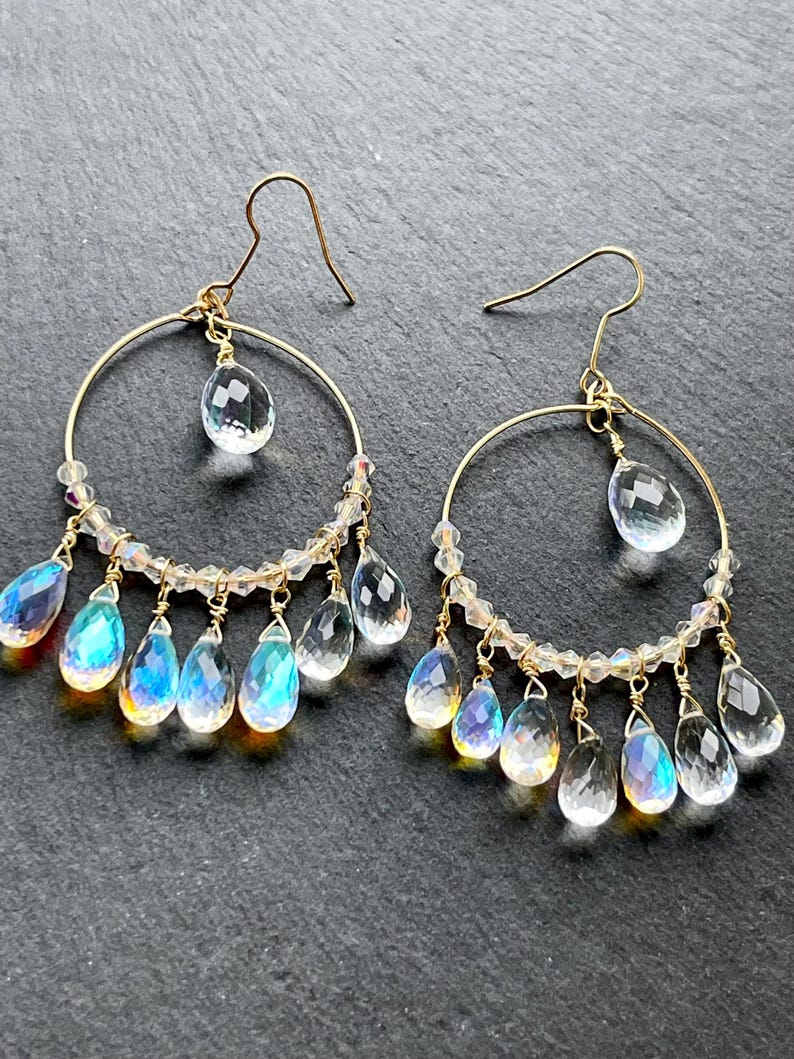 May include: A pair of gold hoop earrings with clear and iridescent teardrop-shaped crystals. The earrings have a delicate design with small clear beads and gold hooks. The crystals reflect a rainbow of colors.