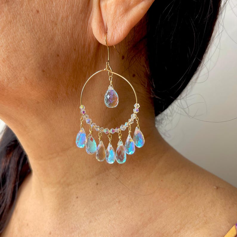 May include: Gold-tone hoop earrings with iridescent teardrop-shaped crystals. The earrings feature a large circular hoop with a smaller teardrop crystal suspended from the top and multiple teardrop crystals dangling from the bottom.