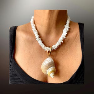 May include: A white shell necklace with a large, spiral-shaped pendant. The necklace features a strand of small, irregular white shell pieces. The pendant is a natural shell with beige and yellow tones, suspended by a gold-colored ring.
