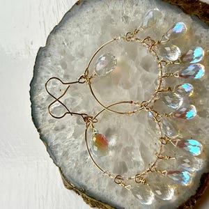 May include: A pair of gold-toned hoop earrings with clear, iridescent teardrop-shaped crystals. The earrings are displayed on a white and gold geode slice, showcasing their delicate design and sparkling details.