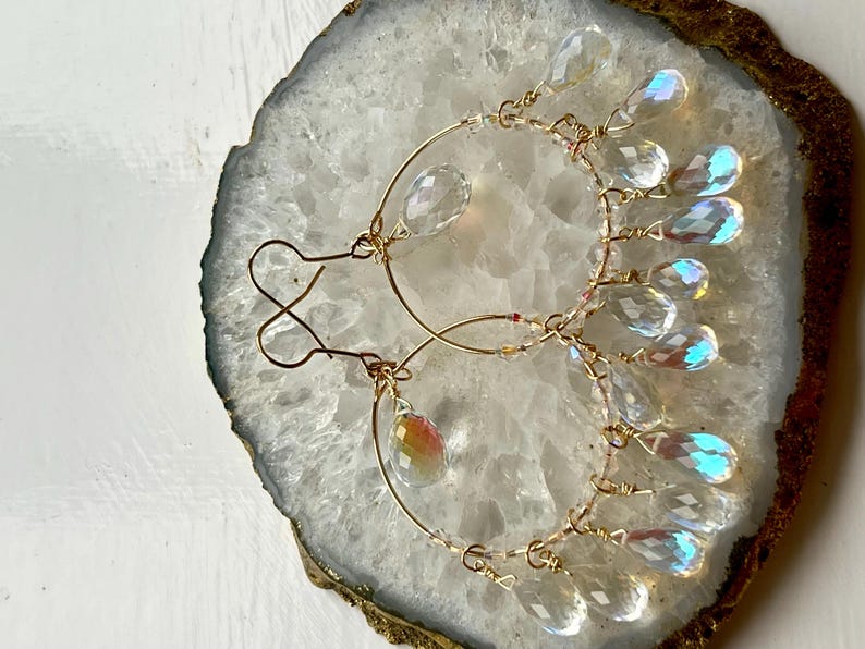 May include: A pair of gold-toned hoop earrings with clear, iridescent teardrop-shaped crystals. The earrings are displayed on a white and gold geode slice, showcasing their delicate design and sparkling details.