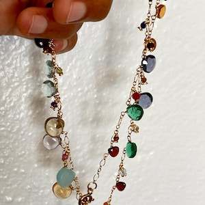 Puede incluir: Un delicado collar de oro con una variedad de piedras preciosas de colores en varias formas y tamaños. Las piedras preciosas incluyen tonos de azul, verde, amarillo y rojo, ensartadas en una fina cadena de oro. El collar se sostiene sobre un fondo blanco.
