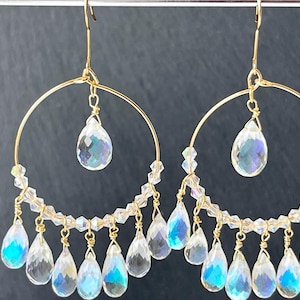May include: Gold-tone hoop earrings with clear, iridescent teardrop-shaped crystals. The earrings feature a delicate design with multiple dangling crystals and smaller beads, creating a shimmering effect. The jewelry is displayed against a dark background.