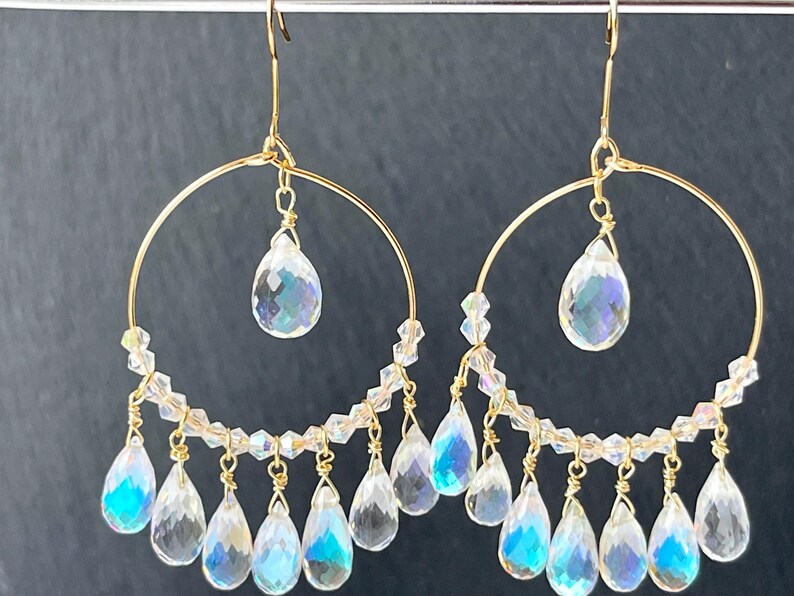 May include: Gold-tone hoop earrings with clear, iridescent teardrop-shaped crystals. The earrings feature a delicate design with multiple dangling crystals and smaller beads, creating a shimmering effect. The jewelry is displayed against a dark background.