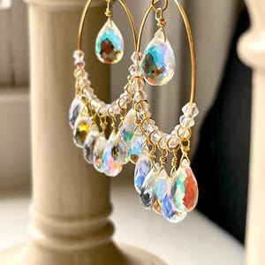 May include: A pair of gold-toned hoop earrings. Each earring features a teardrop-shaped crystal pendant at the top and a cascade of smaller, iridescent crystal droplets. The crystals reflect a rainbow of colors, creating a shimmering effect.