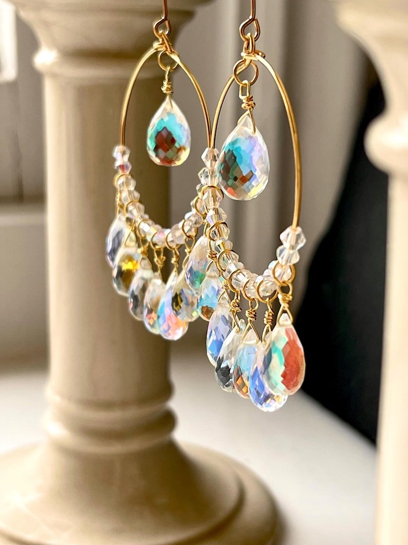 May include: A pair of gold-toned hoop earrings. Each earring features a teardrop-shaped crystal pendant at the top and a cascade of smaller, iridescent crystal droplets. The crystals reflect a rainbow of colors, creating a shimmering effect.