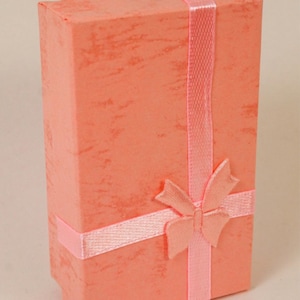 May include: A coral-colored gift box with a textured finish, wrapped with a light pink satin ribbon and a matching bow. The box is rectangular and stands upright, suggesting it is for jewelry or a small gift.