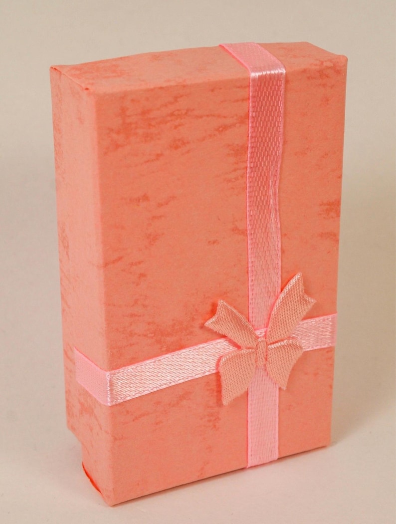 May include: A coral-colored gift box with a textured finish, wrapped with a light pink satin ribbon and a matching bow. The box is rectangular and stands upright, suggesting it is for jewelry or a small gift.