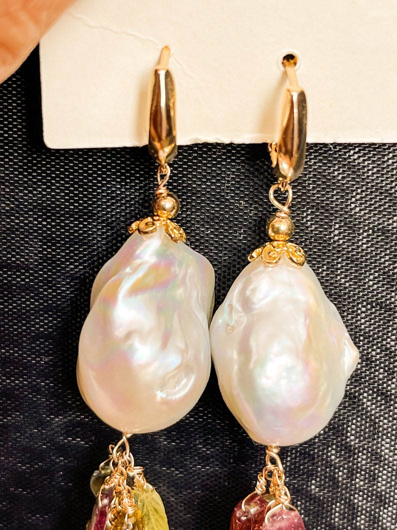 May include: A pair of gold hoop earrings with a large white baroque pearl dangling from each hoop.  Each pearl is accented with a gold filigree design and a cluster of small, colorful gemstones.