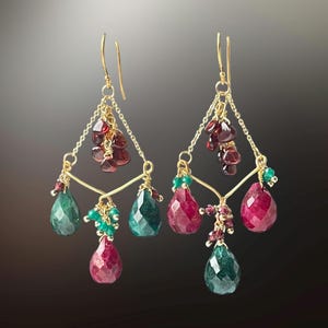 May include: A pair of gold-toned chandelier earrings. Each earring features a triangular design with dangling faceted gemstones in shades of red, green, and gold. Small clusters of red and green beads add detail to the design.