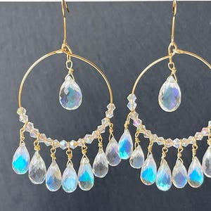 May include: A pair of gold-toned hoop earrings with clear, iridescent teardrop-shaped and faceted crystal pendants. The earrings have a delicate design with smaller clear beads and a larger teardrop crystal suspended from the center of the hoop.