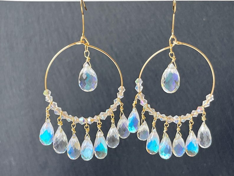 May include: A pair of gold-toned hoop earrings with clear, iridescent teardrop-shaped and faceted crystal pendants. The earrings have a delicate design with smaller clear beads and a larger teardrop crystal suspended from the center of the hoop.