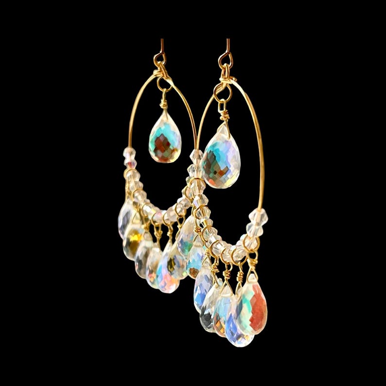 May include: A pair of gold-toned hoop earrings with clear, iridescent teardrop-shaped crystals. Smaller crystals and beads are also incorporated into the design, creating a sparkling effect. The earrings hang against a black background.