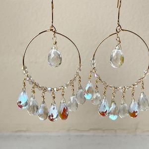May include: A pair of gold-tone hoop earrings with clear, iridescent teardrop-shaped crystals. The earrings feature a large crystal at the top and smaller crystals dangling from the bottom of the hoop. The crystals reflect light, creating a sparkling effect.