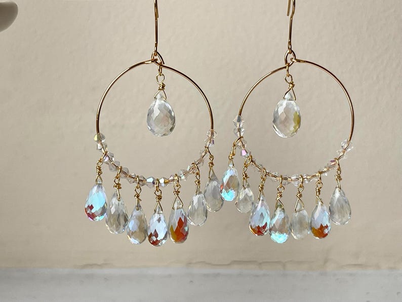 May include: A pair of gold-tone hoop earrings with clear, iridescent teardrop-shaped crystals. The earrings feature a large crystal at the top and smaller crystals dangling from the bottom of the hoop. The crystals reflect light, creating a sparkling effect.