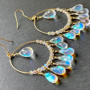 May include: A pair of gold-colored hoop earrings with clear, iridescent teardrop-shaped and round crystal accents. The earrings have a delicate design with multiple crystal dangles, creating a shimmering effect. The hooks are gold-colored.