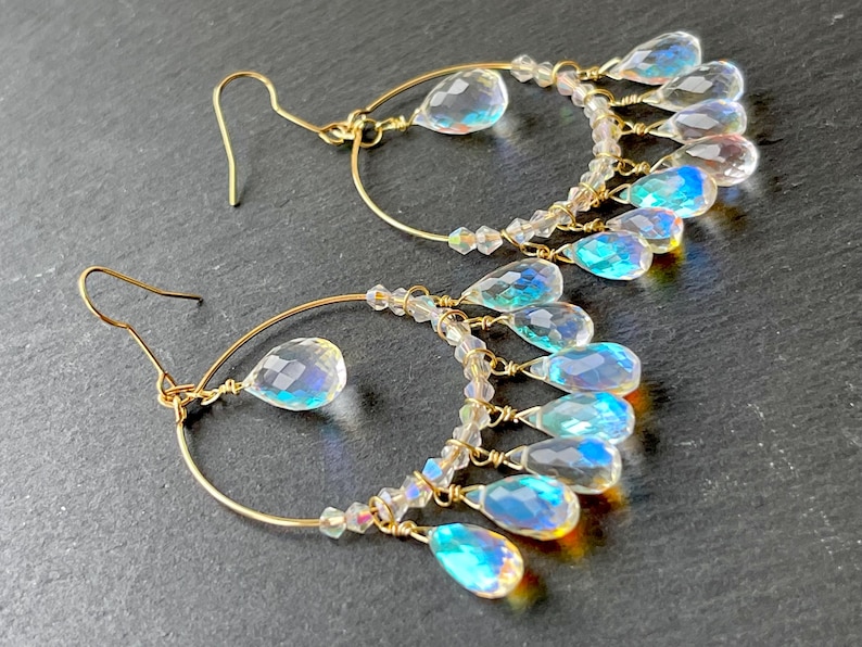 May include: A pair of gold-colored hoop earrings with clear, iridescent teardrop-shaped and round crystal accents. The earrings have a delicate design with multiple crystal dangles, creating a shimmering effect. The hooks are gold-colored.