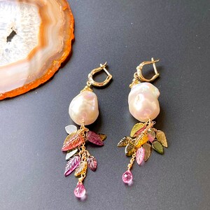 May include: A pair of gold earrings with a large white pearl drop and a dangling charm of green, pink, and brown leaves.