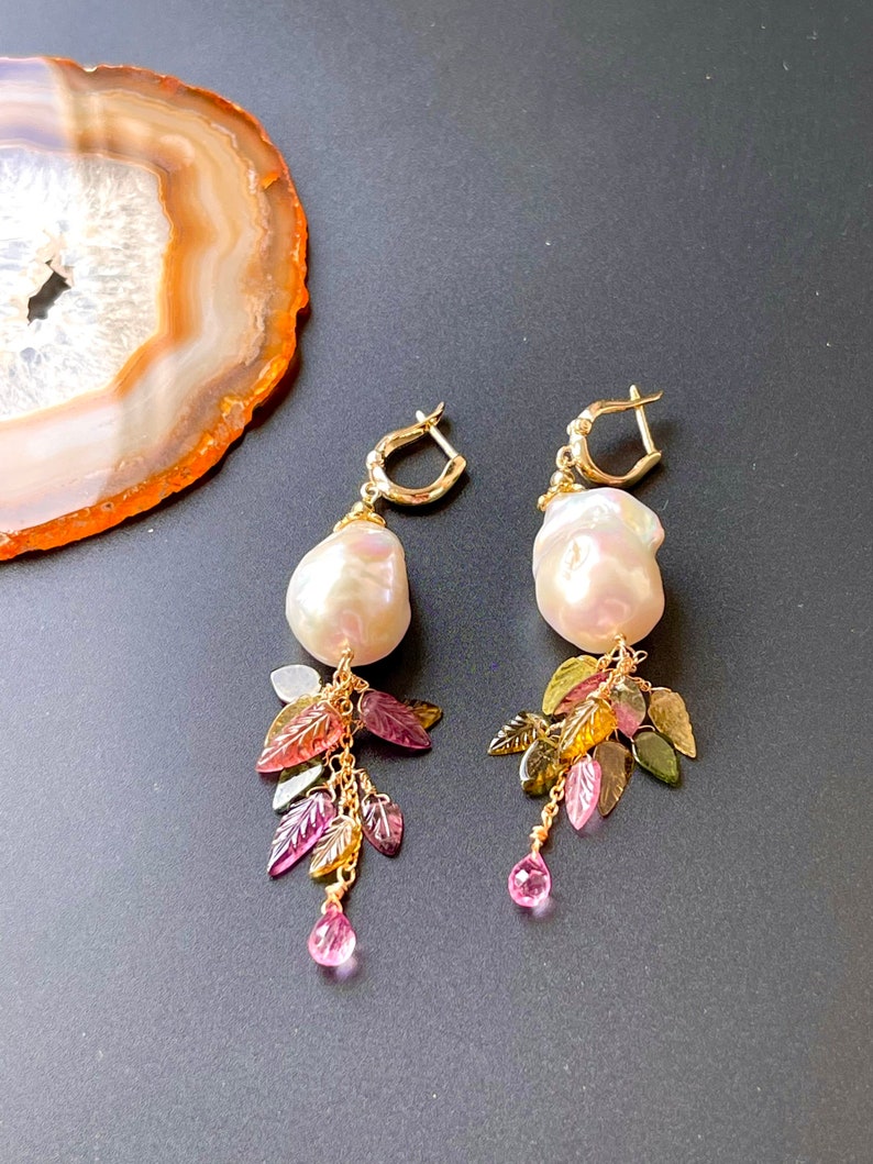 May include: A pair of gold earrings with a large white pearl drop and a dangling charm of green, pink, and brown leaves.