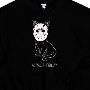 Puede incluir: Sudadera con capucha negra con una ilustración blanca de un gato con una máscara de hockey. El texto "Almost Friday" está impreso debajo del gato.