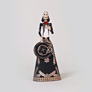 May include: A black and gold Day of the Dead skeleton figurine wearing a traditional Mexican outfit with a red bow tie and a black hat. The figurine is holding a black and gold sombrero.