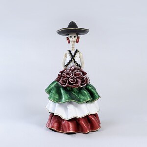Pode incluir: Uma figura de esqueleto do Dia dos Mortos vestindo um sombrero preto, uma blusa branca e uma saia de camadas verde, branca e vermelha com rosas vermelhas.