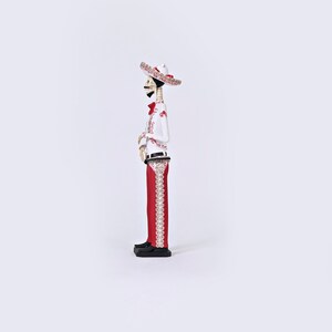 Op de afbeelding: Een wit skeletfiguur dat een rood-witte sombrero draagt, een wit shirt met rode borduursels en een rode broek met witte borduursels. Het figuur houdt een gitaar vast.