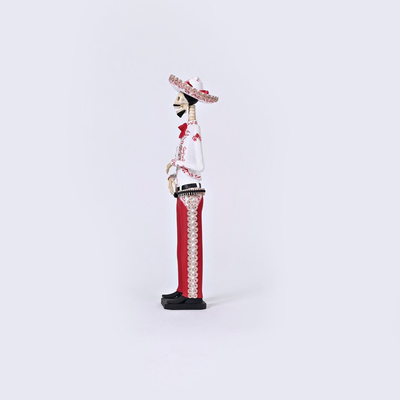 Op de afbeelding: Een wit skeletfiguur dat een rood-witte sombrero draagt, een wit shirt met rode borduursels en een rode broek met witte borduursels. Het figuur houdt een gitaar vast.